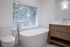 a white bath tub sitting next to a toilet under a bathroom window covered in blinds