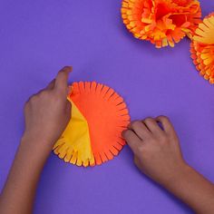 someone making paper flowers out of construction paper on a purple surface with orange and yellow pom - poms