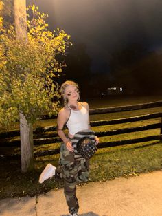 a woman in camo pants and white tank top standing on a skateboard at night