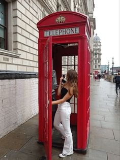 a woman in white pants and black top taking a photo inside a red phone booth