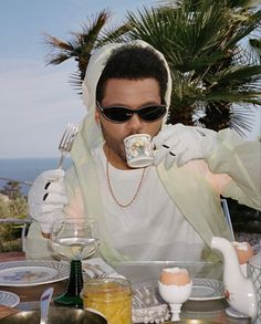 a man wearing sunglasses drinking from a teapot on top of a table with plates and cups