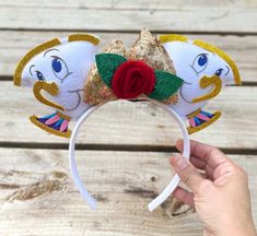 a hand is holding up a white headband with gold sequins and a red rose on it