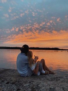 a man and woman sitting on the edge of a body of water watching the sunset