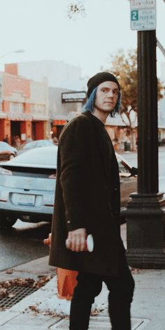 a man with blue hair is walking down the street while wearing a black coat and hat