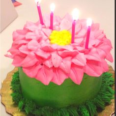 a green cake with pink flowers and lit candles on it's top, sitting on a gold plate