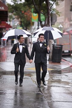 two men in tuxedos walking down the street holding umbrellas