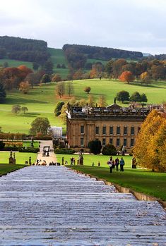 people are walking on the grass in front of a large building with many trees around it