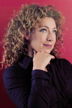 a woman with curly hair and blue eyes posing for a photo in front of a red wall