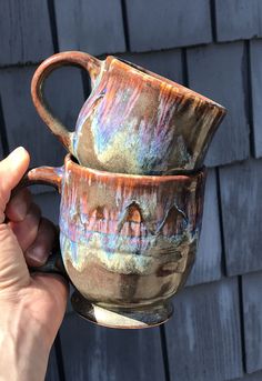 a hand holding two coffee mugs in front of a brick wall with blue and pink paint