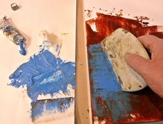 a person is painting with blue and red paint on white paper next to a hand holding a brush