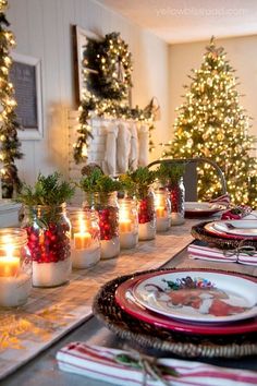 the table is set for christmas dinner with candles in mason jars and decorated trees behind it