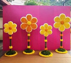 three yellow and orange flowers are in front of a pink wall with grass on it