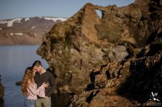 a man and woman standing next to each other near the ocean with mountains in the background