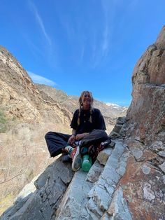 a woman sitting on top of a rocky cliff next to a mountain side under a blue sky