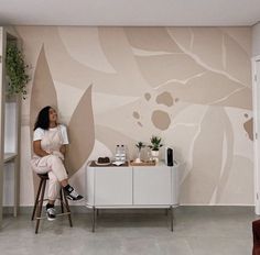 a woman sitting on a stool in front of a wall with flowers painted on it