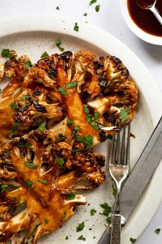 a white plate topped with cooked cauliflower next to a bowl of sauce and a fork