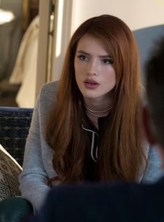 a woman with long red hair sitting on a couch talking to another person in the background