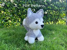 a crocheted gray and white stuffed animal sitting in the grass with bushes behind it