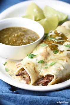 a white plate topped with two burritos next to a bowl of green salsa