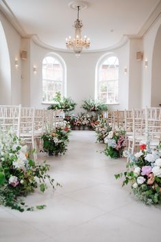 an indoor wedding ceremony with white chairs and floral arrangements on the aisle, chandelier and windows