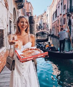 a woman in a white dress is holding a box of pizza and standing on a canal