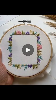 someone is holding up a small embroidery project in front of a white table with flowers on it