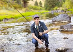 a man kneeling down in the water while holding a fishing rod and wearing a hat