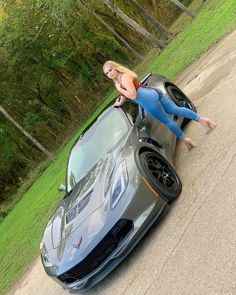 a woman leaning on the hood of a gray sports car with her hand on the wheel