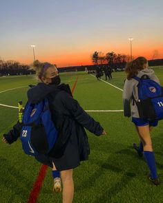 two girls with backpacks walking on a soccer field at sunset or dawn, one carrying a back pack and the other carrying a frisbee