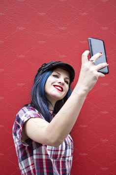 a woman with blue hair taking a selfie on her cell phone while standing in front of a red wall