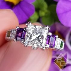 a woman's hand is holding an engagement ring with three stones on it and purple flowers in the background