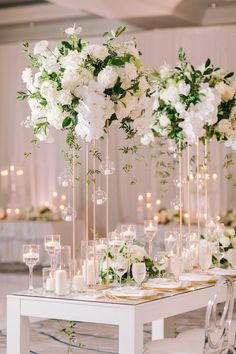 white flowers and greenery are arranged in tall glass vases on a long table