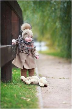 a small child wearing a coat and scarf leaning against a wooden fence with trees in the background