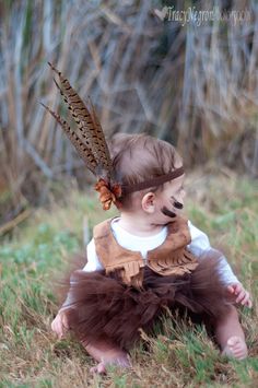 a baby sitting in the grass wearing a brown feather headband with feathers on it