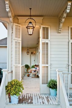 two people sitting on a porch in front of a house with shutters and potted plants