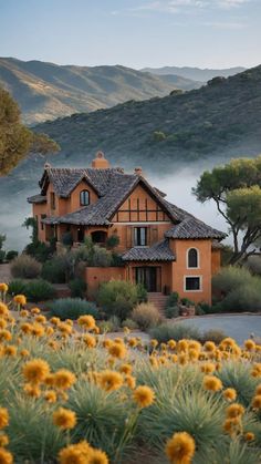 a house with mountains in the background and yellow flowers on the foreground, surrounded by trees