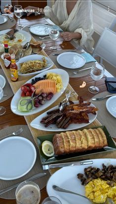 a woman sitting at a table filled with plates and bowls full of food, including eggs, sausages, toast, fruit, and bread