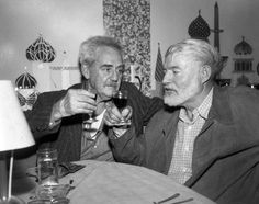 two older men sitting at a table with wine glasses in their hands and one holding a drink