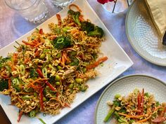 a white plate topped with noodles and veggies next to another plate filled with broccoli