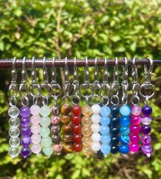 a bunch of different colored beads hanging from a metal hook on a tree branch in front of some bushes