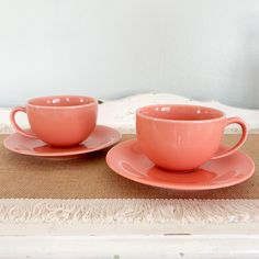 two pink cups and saucers sitting on top of a placemat next to each other