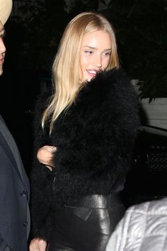 the blonde woman is wearing black leather pants and a fur coat as she walks past a man in a suit