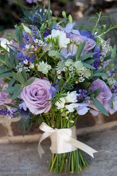 a bouquet of purple and white flowers with greenery on the stems, tied to a stone wall