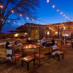 people are sitting around an outdoor fire pit at night with string lights strung across the roof