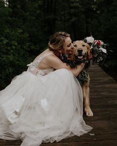 a woman in a wedding dress hugging a dog on a wooden platform with greenery around her