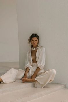 a woman is sitting on the stairs in white clothing and pearls around her neck, looking at the camera