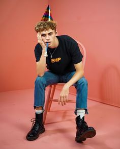 a young man sitting on a chair wearing a party hat and holding a pencil in his mouth