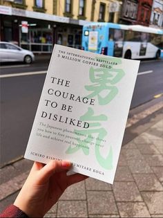 a person holding up a book on the side of a road with buildings in the background