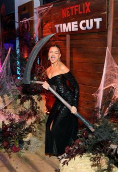 a woman in black dress holding a large knife and posing for the camera with halloween decorations behind her