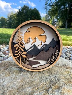 a wooden plaque with mountains and trees on it, sitting on top of a rock